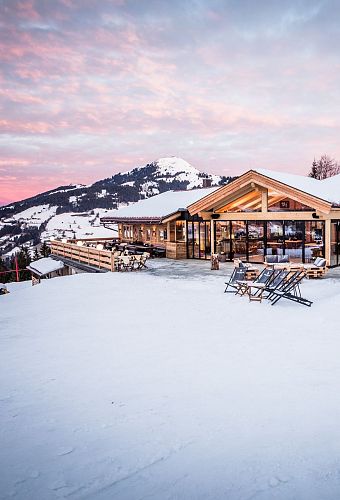 Een moderne berghut met grote ramen en sneeuw. Ligstoelen staan buiten op het terras met een berg op de achtergrond onder een lichtroze hemel.