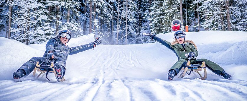 Two people sledding down a snowy slope in a forest, smiling and raising their arms, surrounded by snow-covered trees.