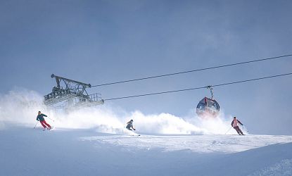 A skier descends a snowy slope under a clear blue sky beside a gondola lift transporting passengers uphill.