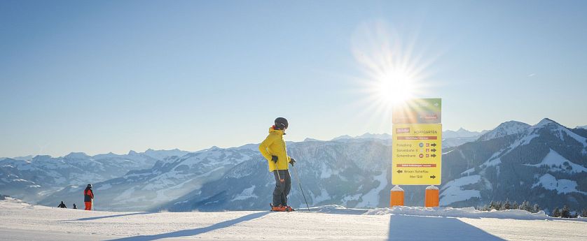 Person in yellow jacket standing on a snowy mountain with ski poles, mountains in the background, and the sun shining brightly overhead.