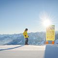 Een skiër in een gele jas naast een wegwijzer op een zonnige bergtop met besneeuwde bergen op de achtergrond.