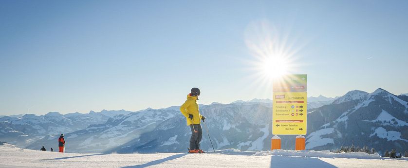 Person in gelber Jacke auf Skiern betrachtet ein Schild im Schnee, umgeben von schneebedeckten Bergen und strahlendem Sonnenschein.