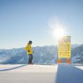 Person in gelber Jacke auf Skiern betrachtet ein Schild im Schnee, umgeben von schneebedeckten Bergen und strahlendem Sonnenschein.