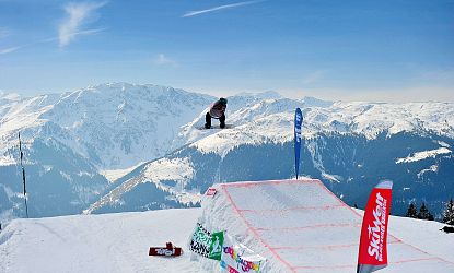 Een snowboarder springt van een schans in een besneeuwd berglandschap met blauwe luchten.