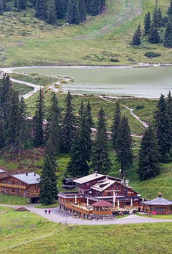 Ein traditionelles Berghaus mit Holzfassade liegt inmitten von grasbewachsenen Hügeln und Tannenbäumen. Wanderwege schlängeln sich durch die Landschaft.