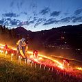 Menschen plaatsen kaarsen op een berghelling tijdens een festival, met bergen in de achtergrond en een dorp onder een bewolkte avondlucht.