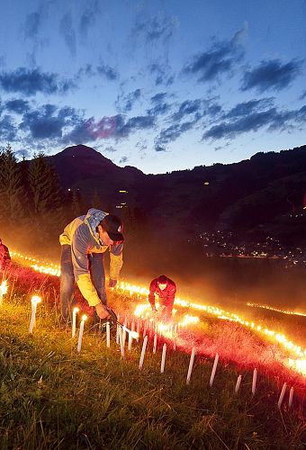 Menschen zünden bei Dämmerung in einer Berglandschaft zahlreiche Kerzen oder Fackeln an, um ein Feuerbild zu formen. Der Himmel ist wolkig-blau.