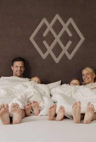 Een lachende familie van vier ligt in bed, voeten uitstrekken onder de dekens. Achtergrond toont een bruin wandpaneel met logo.