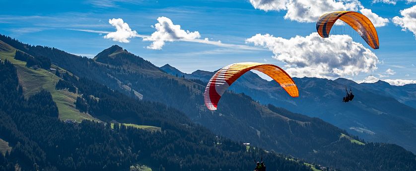 A paraglider with a red and orange canopy flies over a scenic mountain range under a blue sky with fluffy white clouds.