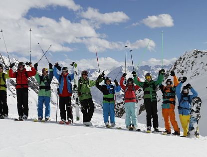 Group of skiers in colorful outfits stand with raised poles on a snowy mountain, showcasing a background of majestic peaks under a partly cloudy sky.