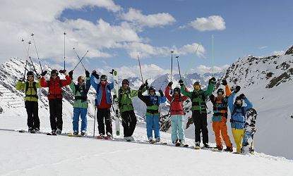 Groep skiërs in kleurrijke jassen op een besneeuwde berg, poseren met skistokken. Bergen en blauwe lucht op de achtergrond, vreugdevolle sfeer.