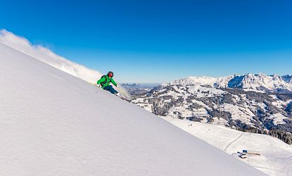 Skiër in groene outfit daalt een besneeuwde berg af onder een heldere blauwe hemel met bergketens op de achtergrond.