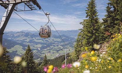 Eine Seilbahnkabine schwebt über einer grünen, hügeligen Landschaft mit Bergen im Hintergrund. Es ist ein klarer, sonniger Tag.