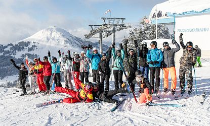 Group of skiers posing happily on a snowy slope with ski lift, wearing colorful outfits and holding skis, against a backdrop of snow-covered mountains.