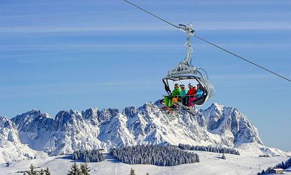 Eine Gruppe von Skifahrern sitzt in bunten Outfits auf einem Sessellift hoch über schneebedeckten Bergen. Der Himmel ist klar und blau, die Berglandschaft spektakulär.