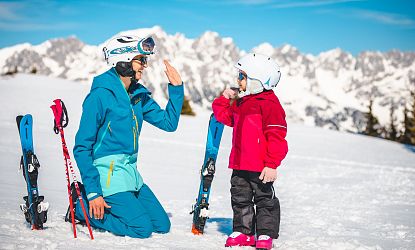 Volwassene en kind high-fiven op sneeuw, dragen ski-uitrusting en helmen, met bergen op de achtergrond.