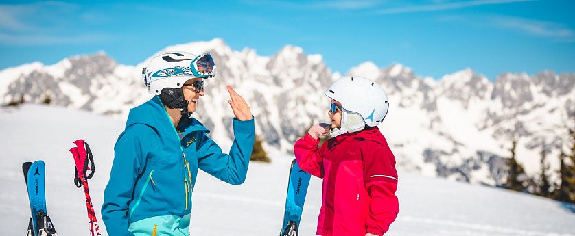 kind-macht-high-five-mit-elternteil-auf-piste-mit-wilder-kaiser-im-hintergrund-2