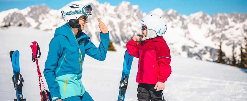 An adult and child in ski outfits high-fiving in a snowy mountain setting. The child is holding skis, both wearing helmets and goggles, with bright attire.