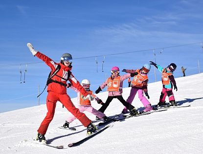 Group of children and an instructor skiing on a sunny slope, all wearing colorful ski gear and helmets, posing energetically for the camera.