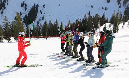 Een groep skiërs staat op een besneeuwde piste in de bergen met hun skistokken omhoog en omringd door bomen en een net, klaar om te starten.