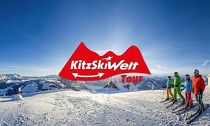 Mehrere Skifahrer stehen auf einem verschneiten Berggipfel mit weitem Panorama. Logo "KitzSkiWelt Tour" ist prominent im Vordergrund.