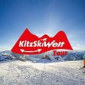 Mehrere Skifahrer stehen auf einem verschneiten Berggipfel mit weitem Panorama. Logo "KitzSkiWelt Tour" ist prominent im Vordergrund.