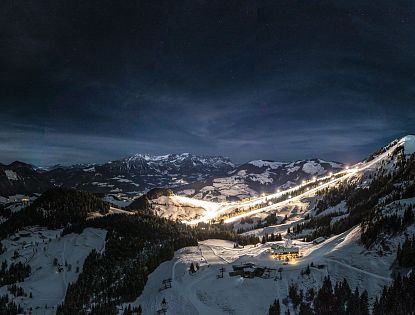Night view of snow-covered mountains with ski slope lights and village under a starry sky.