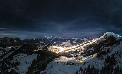 Night view of snow-covered mountains with ski slope lights and village under a starry sky.