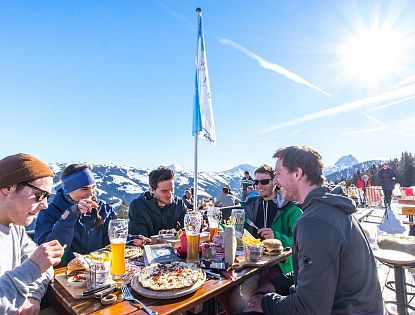 Groep mensen geniet van eten en drinken op een zonovergoten bergterras met uitzicht op besneeuwde bergen en blauwe lucht.