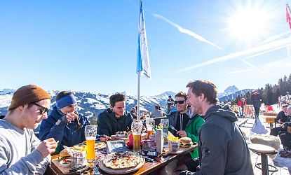 Groep mensen geniet van eten en drinken op een zonovergoten bergterras met uitzicht op besneeuwde bergen en blauwe lucht.