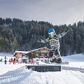 Child wearing a blue helmet and colorful winter gear snowboards on a snowy slope with pine trees and a chalet in the background.