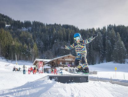 Kind snowboardt op een rail in een besneeuwd skigebied, met heldere kleding en blauwe helm, omgeven door bomen en een houten lodge.