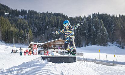 Child wearing a blue helmet and colorful winter gear snowboards on a snowy slope with pine trees and a chalet in the background.