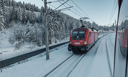 Ein roter Zug fährt auf schneebedeckten Gleisen durch eine winterliche Landschaft, umgeben von verschneiten Tannen. Stromleitungen erstrecken sich über die Strecke.