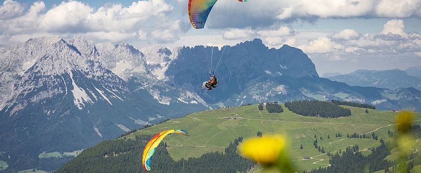 Paragleiter fliegen über grüne Berge mit schneebedeckten Gipfeln, blauer Himmel und Wolken im Hintergrund. Eine Sommerlandschaft in den Alpen.