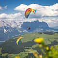 Paragleiter fliegen über grüne Berge mit schneebedeckten Gipfeln, blauer Himmel und Wolken im Hintergrund. Eine Sommerlandschaft in den Alpen.