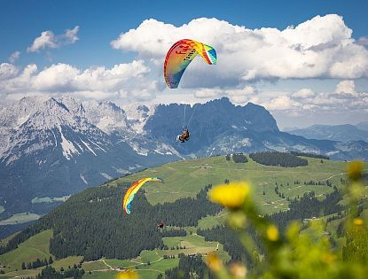 Paragleiter fliegen über grüne Berge mit schneebedeckten Gipfeln, blauer Himmel und Wolken im Hintergrund. Eine Sommerlandschaft in den Alpen.