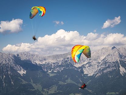 Zwei Paraglider schweben über einer Berglandschaft mit grünen Hügeln und teils bewölktem Himmel, ihre Schirme sind bunt und auffällig.