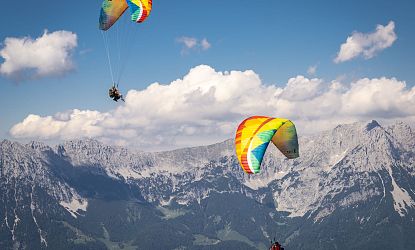 Zwei Paraglider schweben über einer Berglandschaft mit grünen Hügeln und teils bewölktem Himmel, ihre Schirme sind bunt und auffällig.