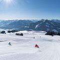 Twee skiërs dalen een besneeuwde berg af onder een heldere lucht, omgeven door bergen en bomen in de verte.