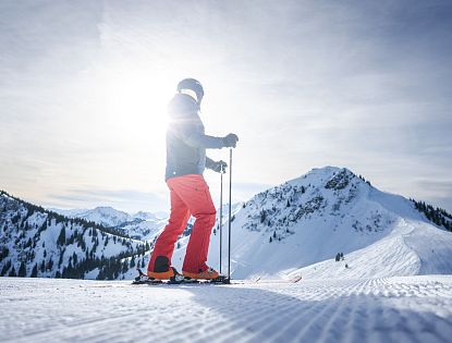 Skifahrer in roter Hose und schwarzer Jacke steht bei Sonnenschein auf einer schneebedeckten Bergspitze mit blauen Himmel im Hintergrund.
