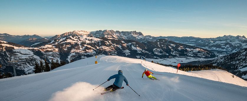 Zwei Skifahrer fahren dynamisch eine schneebedeckte Abfahrt hinunter, umgeben von beeindruckenden Gebirgslandschaften unter blauem Himmel.