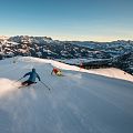 Zwei Skifahrer fahren dynamisch eine schneebedeckte Abfahrt hinunter, umgeben von beeindruckenden Gebirgslandschaften unter blauem Himmel.