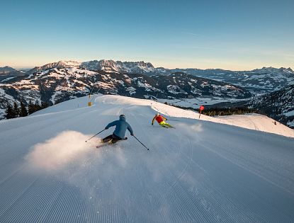 Zwei Skifahrer fahren dynamisch eine schneebedeckte Abfahrt hinunter, umgeben von beeindruckenden Gebirgslandschaften unter blauem Himmel.