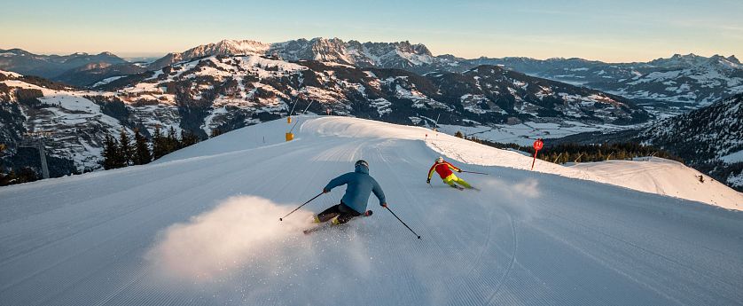 Zwei Skifahrer fahren eine schneebedeckte Piste hinunter, umgeben von majestätischen Bergen unter blauem Himmel. Die Sonne beleuchtet die Winterszene.