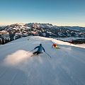 Twee skiërs glijden snel een besneeuwde piste af tegen een achtergrond van imposante bergen bij zonsopgang, terwijl sneeuw opwaait.