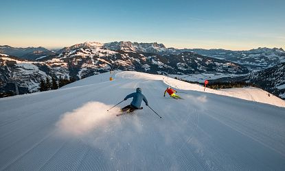Twee skiërs glijden snel een besneeuwde piste af tegen een achtergrond van imposante bergen bij zonsopgang, terwijl sneeuw opwaait.