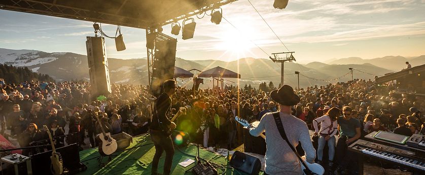 Musicians play on stage at an outdoor festival with mountains and a sunset, surrounded by a lively crowd enjoying the performance.