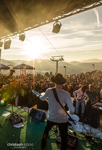 Eine Band spielt auf einer Open-Air-Bühne in einer Berglandschaft bei Sonnenuntergang. Viele Zuschauer genießen die Musik im Freien.