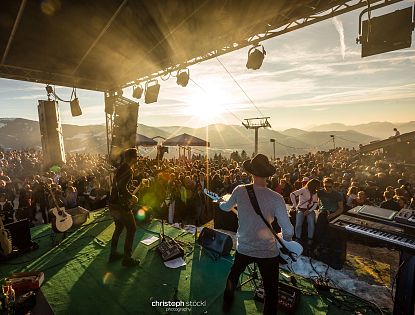 Band speelt op buitenpodium in de bergen bij zonsondergang, omringd door groot publiek en bergachtergrond.